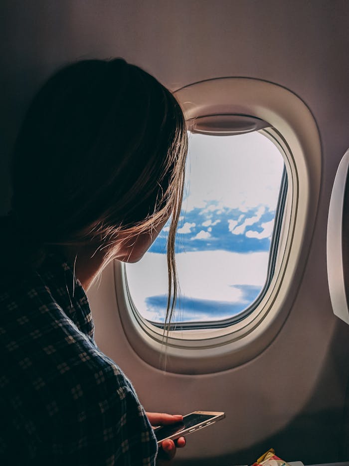 services-01 A woman looks out an airplane window, capturing a serene view of clouds and sky during a flight.